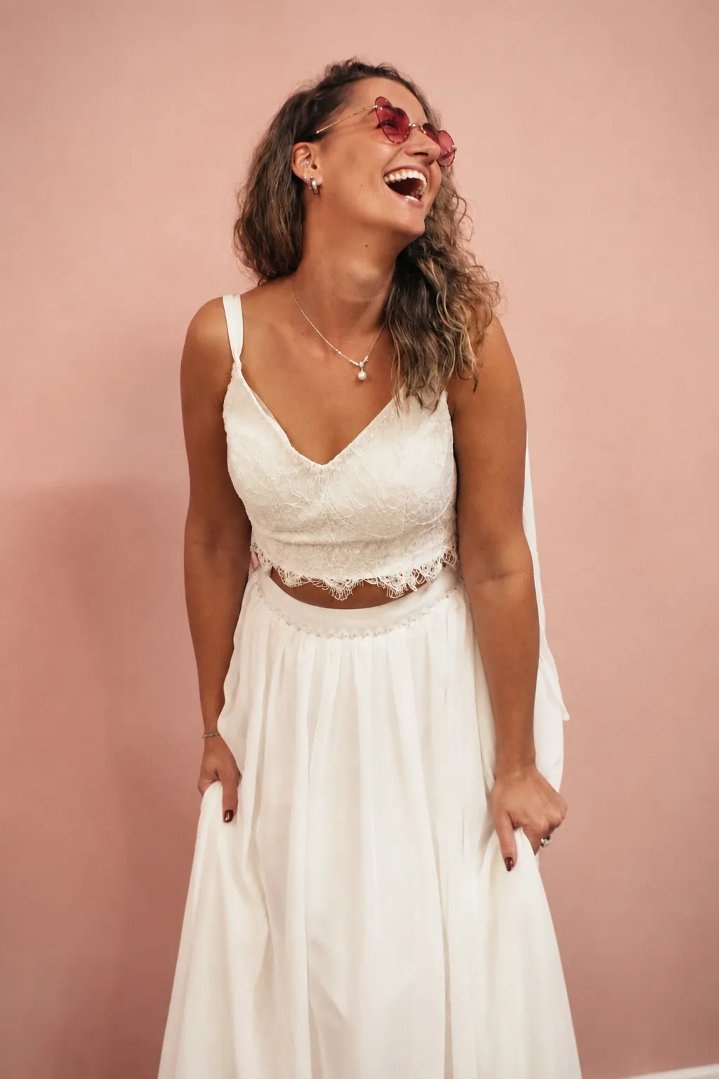 Lachende bruid in een tweedelige boho trouwjurk tegen de roze wand
